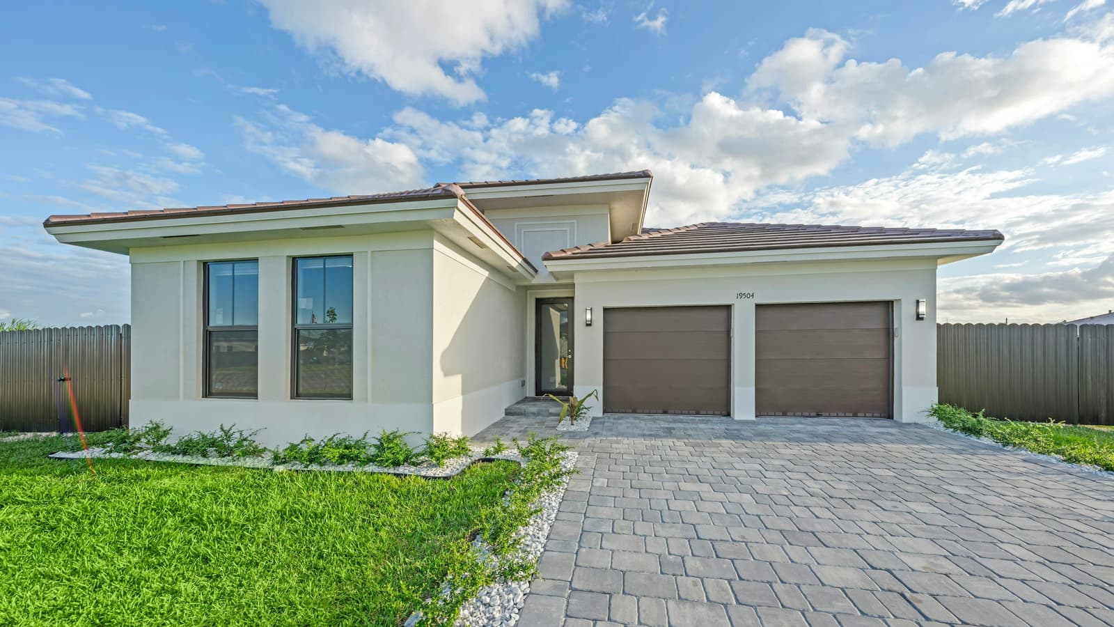 Modern single-story stucco home with two-car garage, paver driveway and green lawn under a blue sky